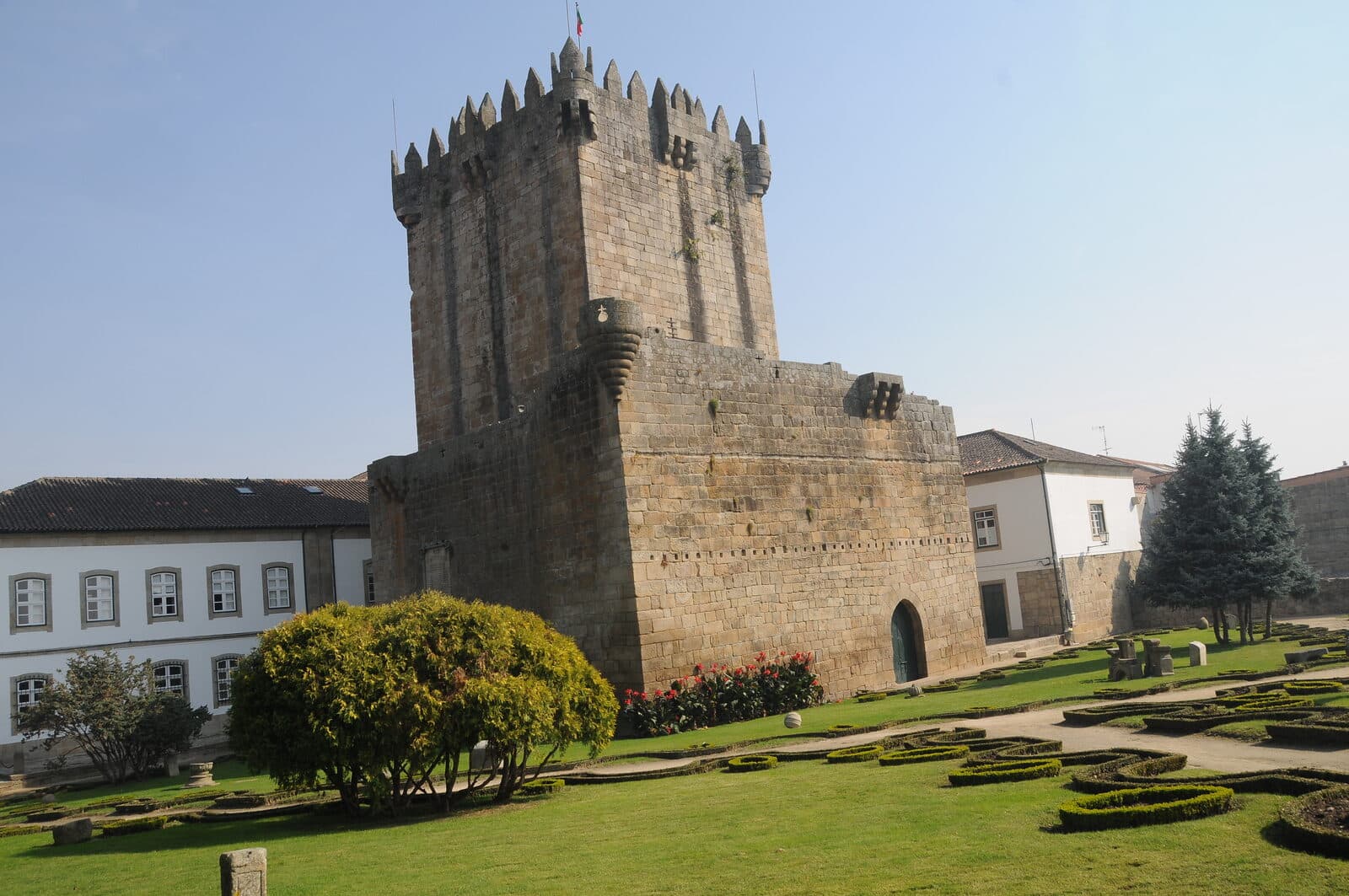 Torre de menagem do castelo de Chaves, de granito e com ameias, rodeada por um jardim com canteiros e casas brancas