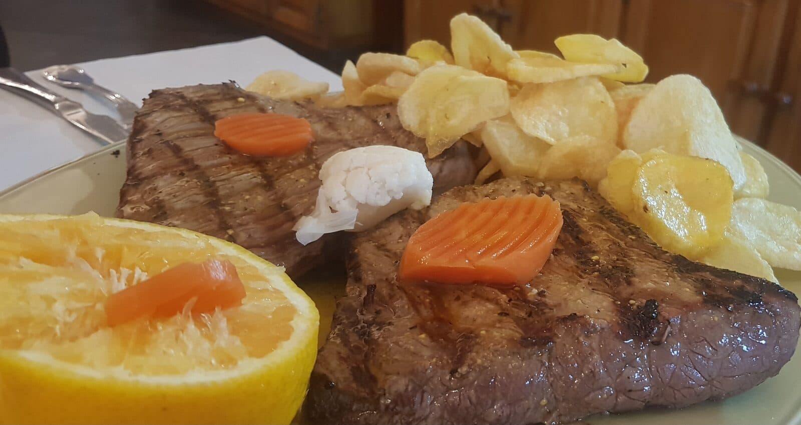 Duas postas de carne de vaca grelhadas, com as marcas da grelha, servidas com batata frita às rodelas, cenoura e meia laranja