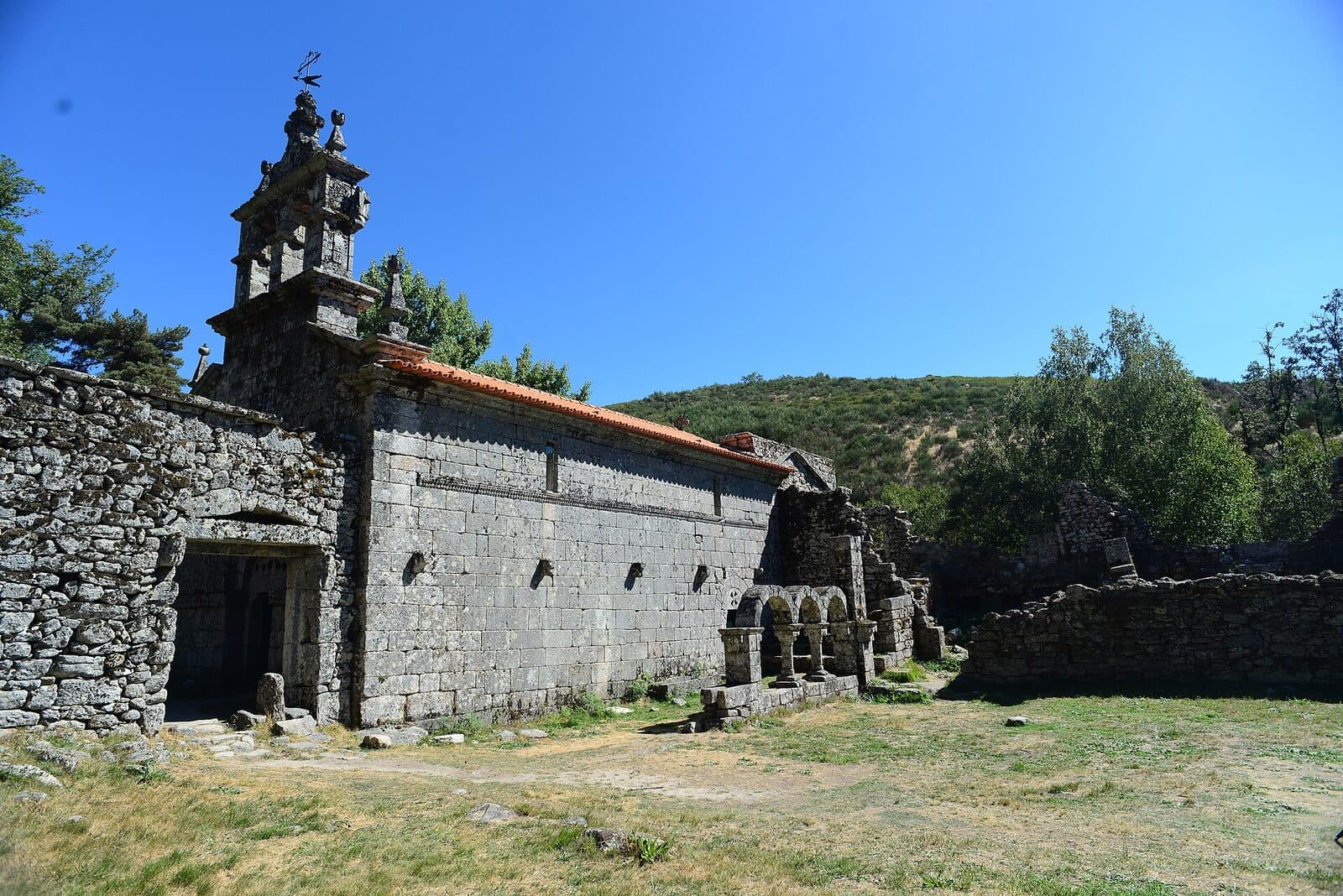 Igreja do Mosteiro de Santa Maria das Júnias, em granito, com campanário e arcos do antigo claustro, num vale verde perto de Pitões das Júnias