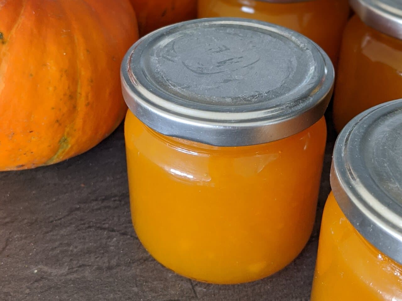 Frascos de doce de abóbora em cima de uma laje escura, com duas abóboras cor de laranja ao lado