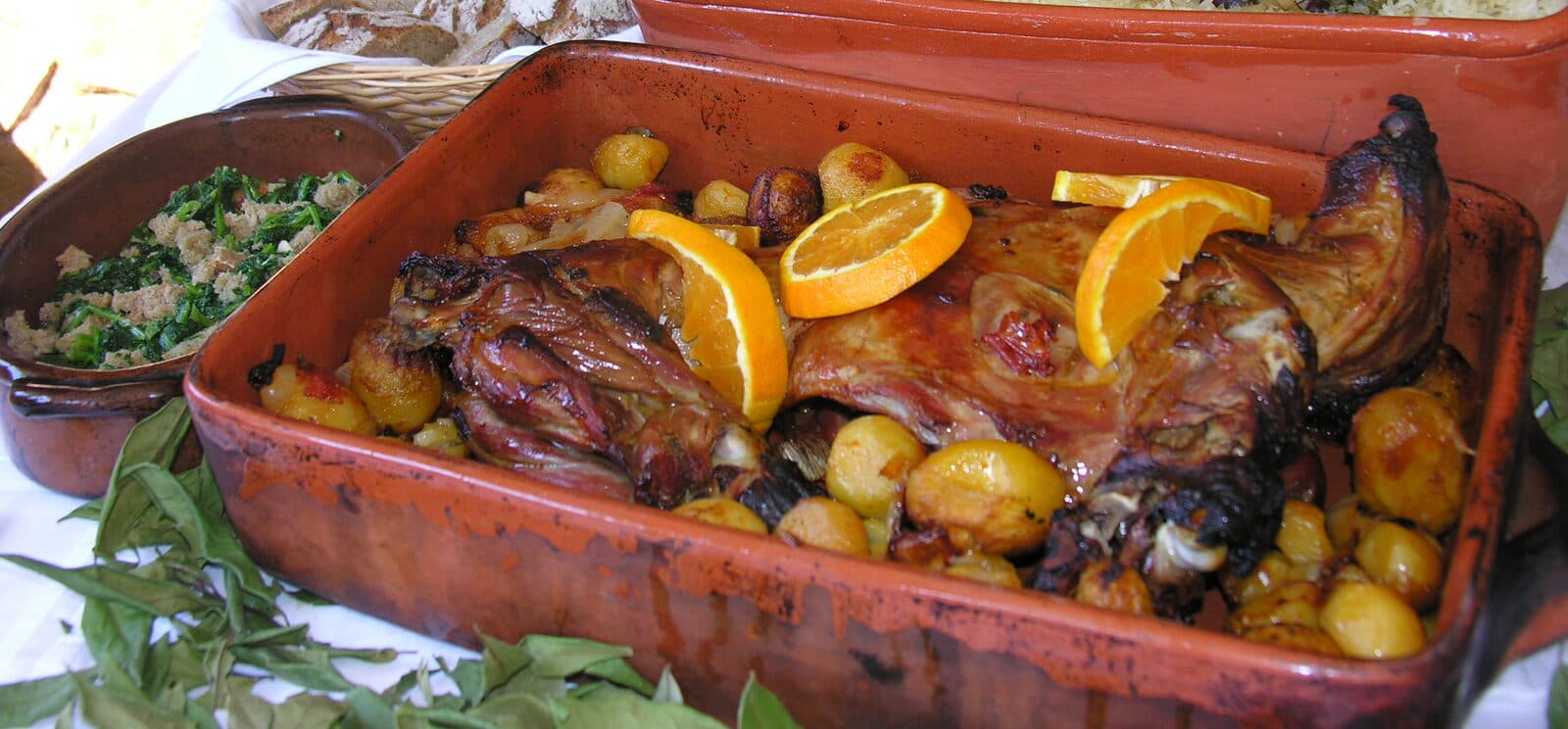 Cabrito assado com batatas pequenas e rodelas de laranja, num tabuleiro de barro