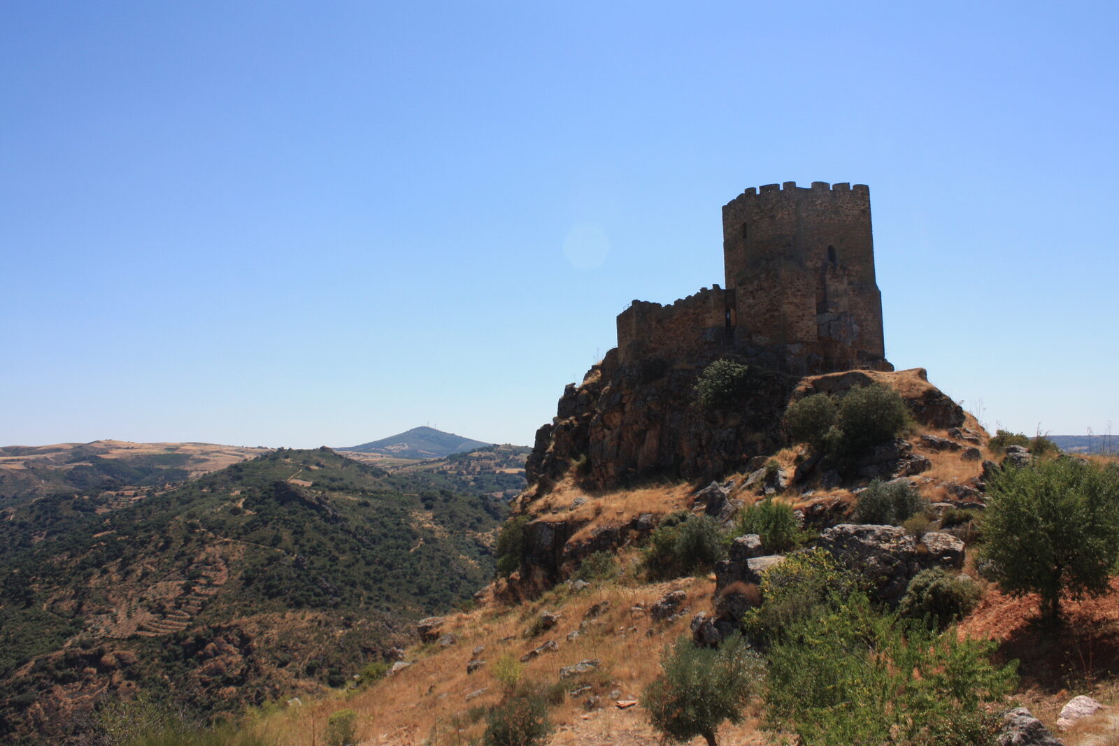 Torre de menagem e muralha do castelo de Algoso no alto de um penhasco, sobre encostas secas com oliveiras e um vale ao fundo