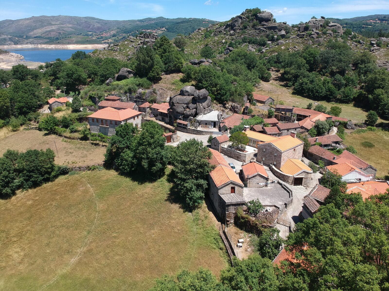 Vista aérea de Sirvozelo: casas de granito com telhados de telha entre grandes penedos e carvalhos, com a albufeira da Paradela ao fundo