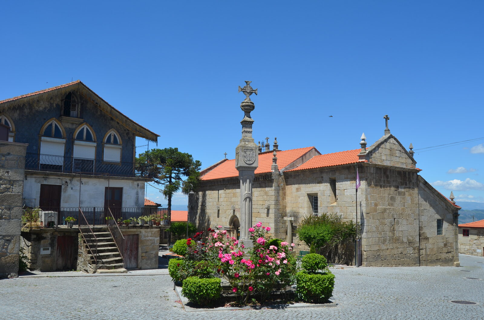 Largo empedrado de Barcos com um cruzeiro de granito no meio de um canteiro de rosas, uma igreja de granito com telhado de telha e uma casa azulejada à esquerda