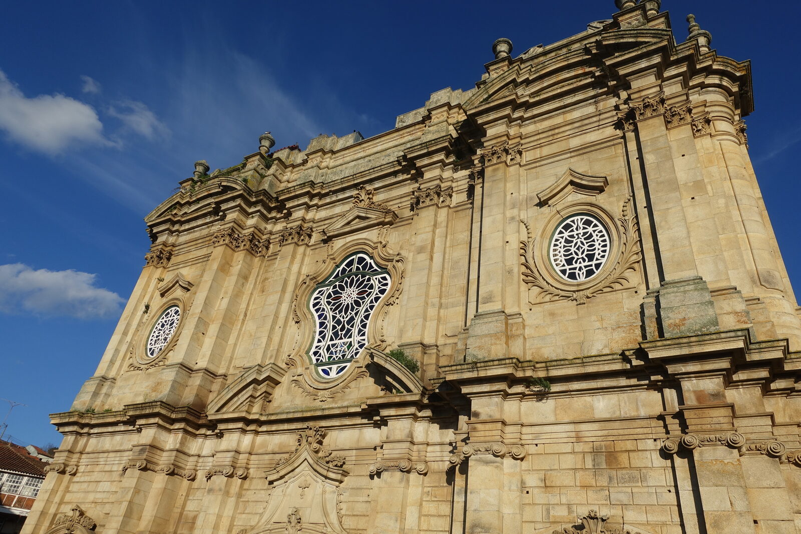 Fachada barroca de granito dourado da igreja do mosteiro de Salzedas, vista de baixo, com uma janela recortada com vitral e dois óculos, contra um céu azul