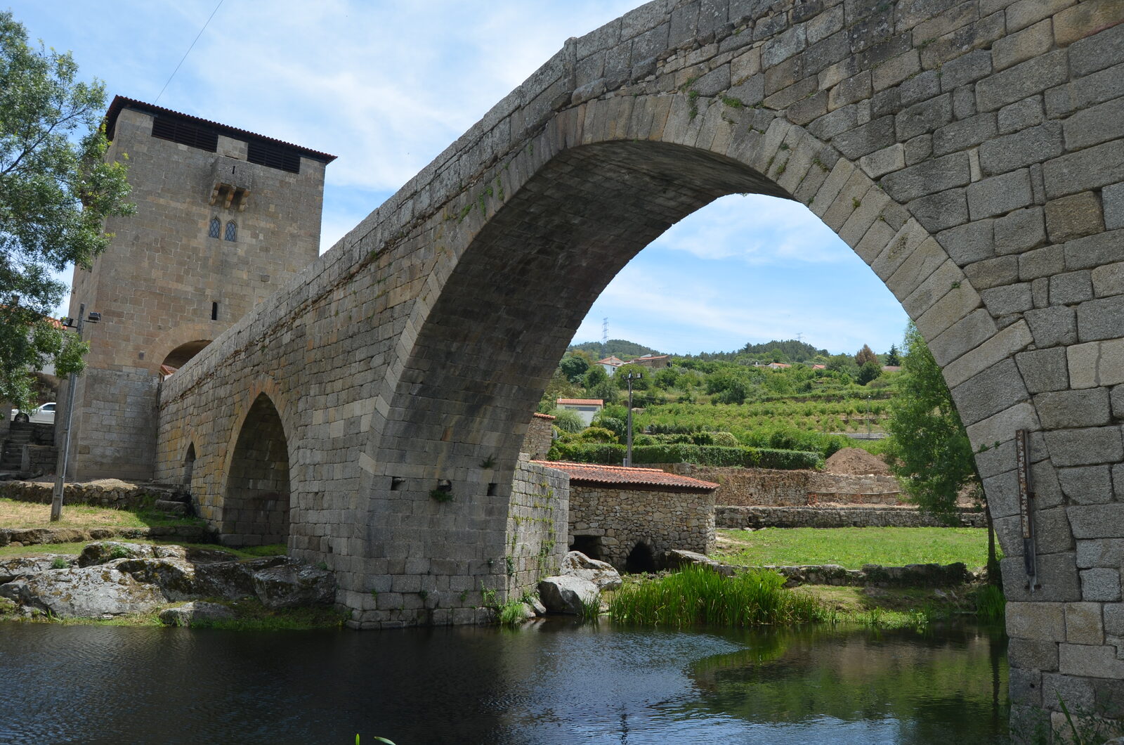 Ponte de granito com um grande arco em ogiva sobre o rio Varosa, com a torre quadrada de Ucanha à esquerda e socalcos verdes ao fundo