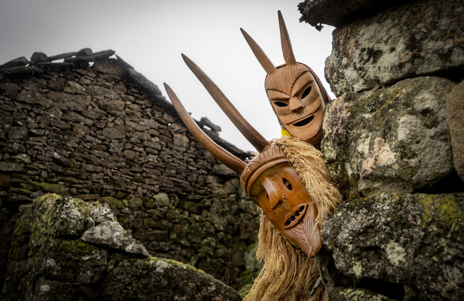 Dois caretos de Lazarim com máscaras de madeira de cornos longos e expressão demoníaca, a espreitar por trás de um muro de pedra