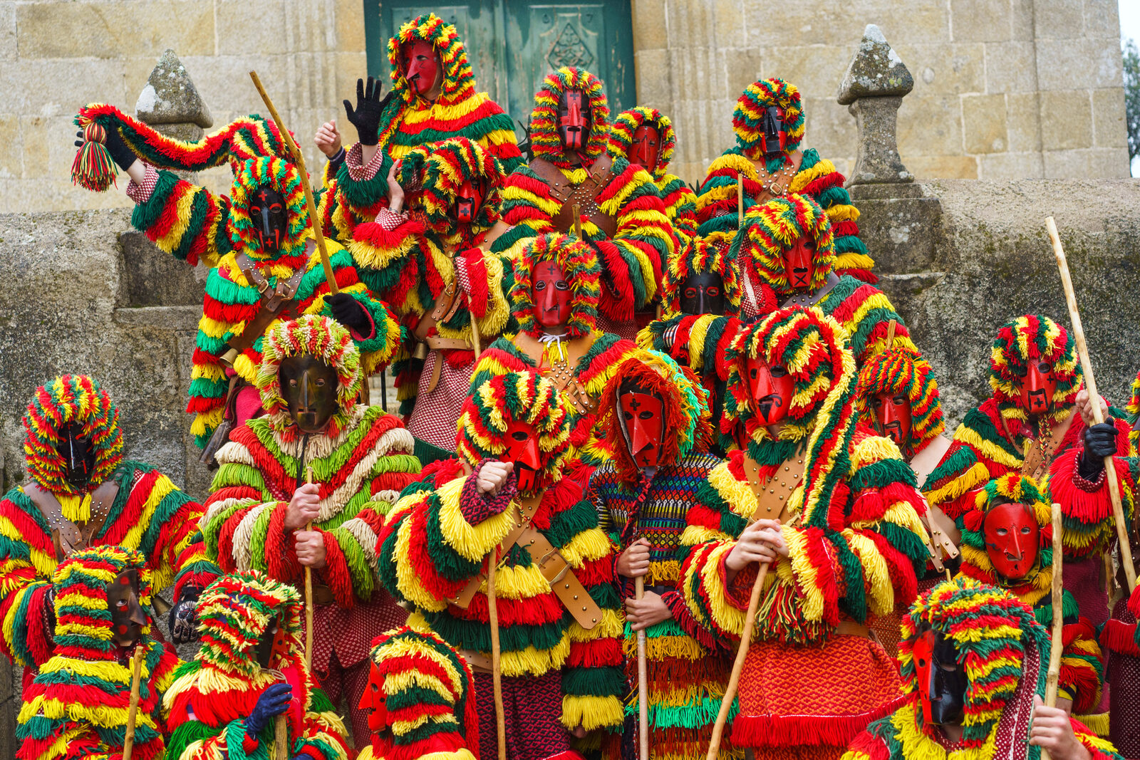 Grupo de caretos de Podence com máscaras vermelhas e pretas e fatos de franjas vermelhas, amarelas e verdes, na escadaria de uma igreja de granito