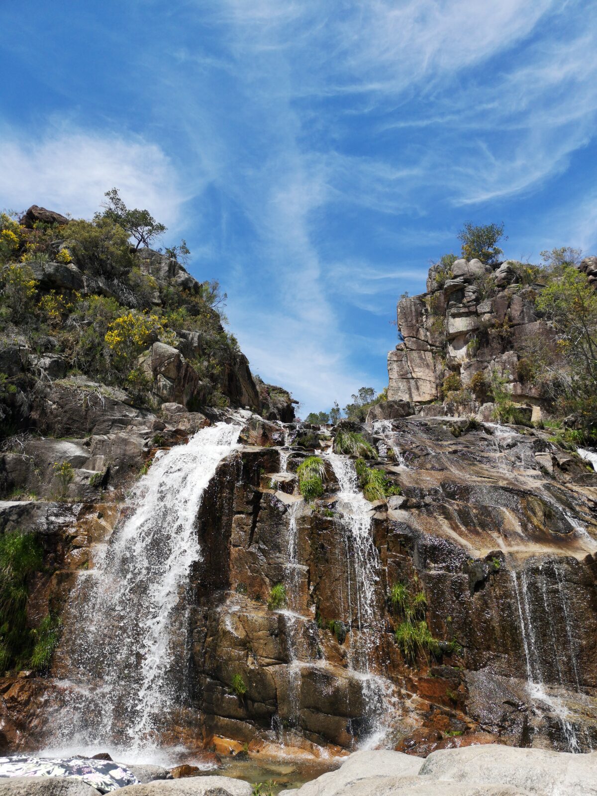 Água a cair em dois braços por degraus de granito escuro, com céu azul e nuvens altas
