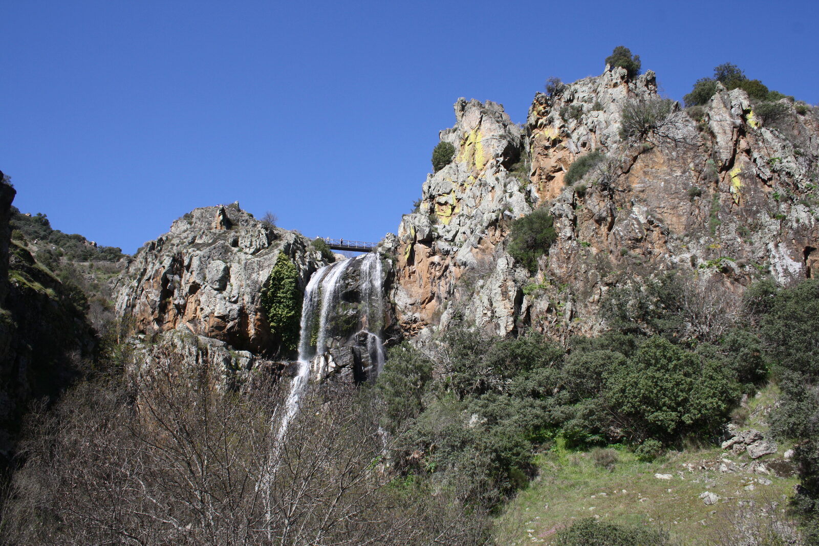 Cascata a cair entre fragas de rocha clara e alaranjada, com uma ponte metálica no alto e céu azul