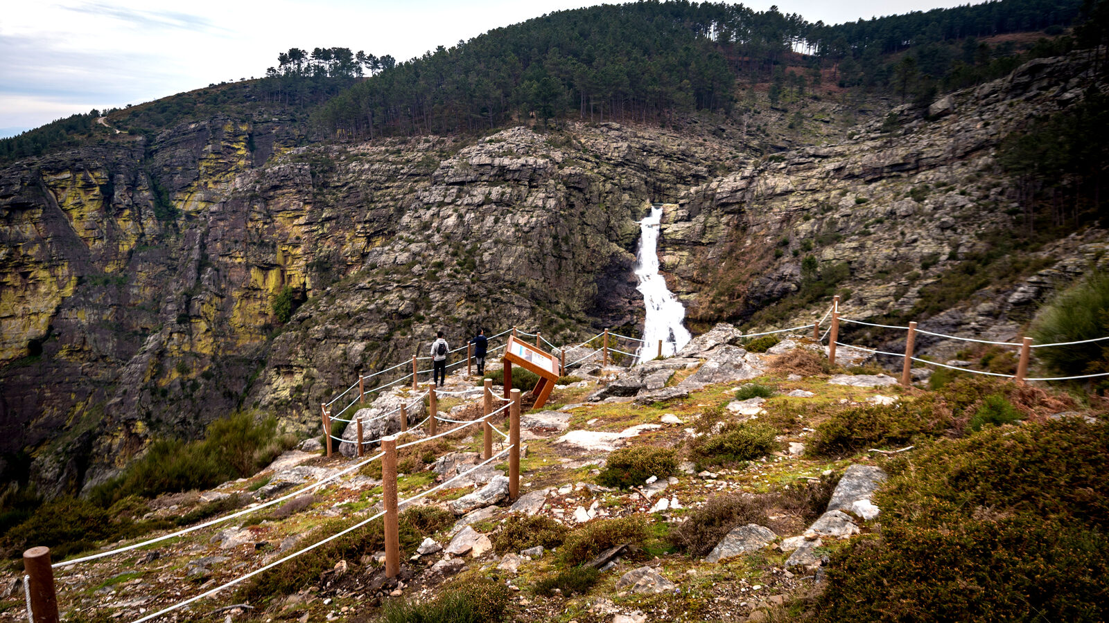 Quedas brancas do rio Olo numa escarpa de rocha, vistas de um caminho com vedação de corda e duas pessoas ao longe