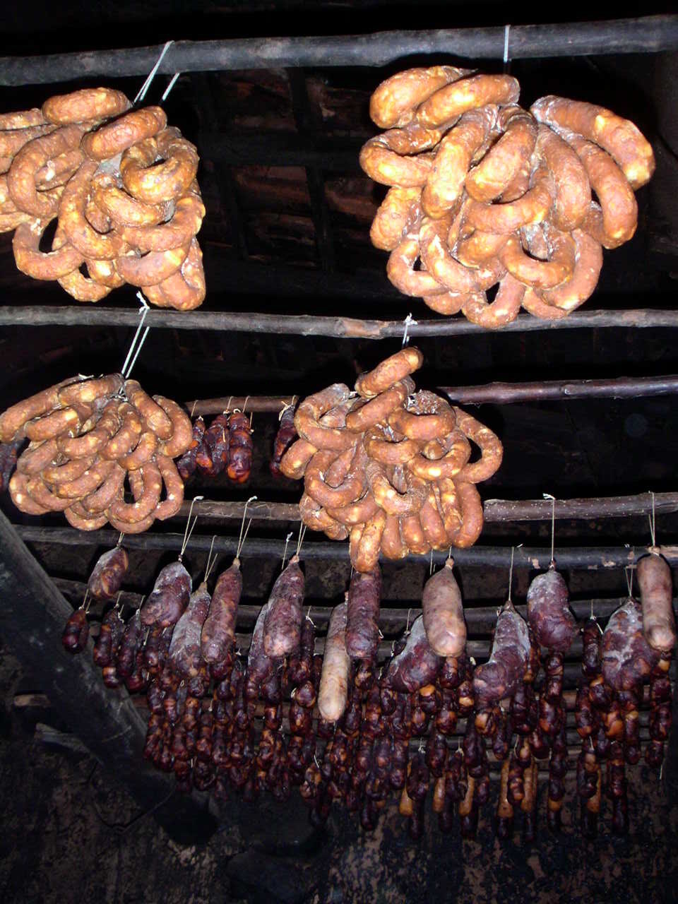 Chouriças e alheiras penduradas em varas por cima de uma lareira, numa cozinha antiga