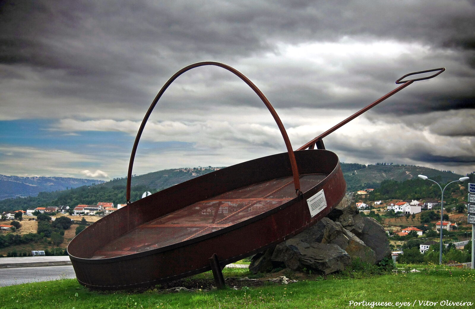 Assador de castanhas gigante em ferro, pousado sobre pedras num relvado, com a vila de Vinhais e os montes ao fundo