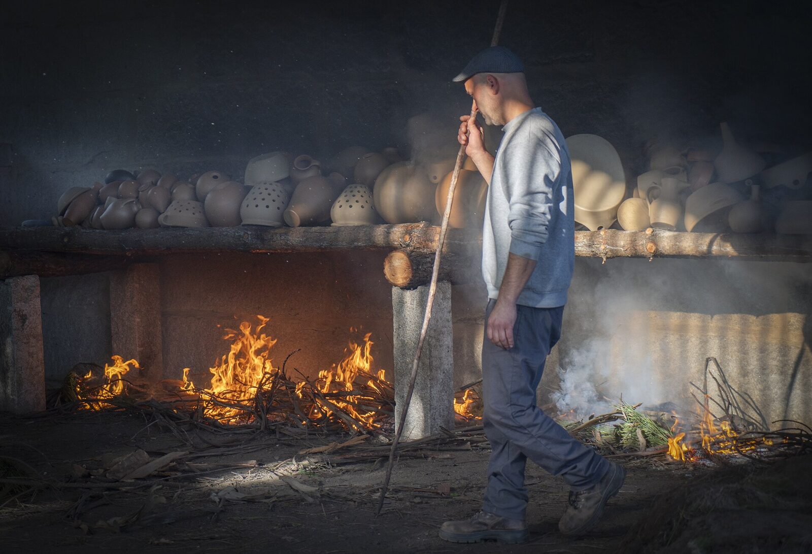 Oleiro junto a um forno com lume aceso e prateleiras com peças de barro, na primeira cozedura de louça preta