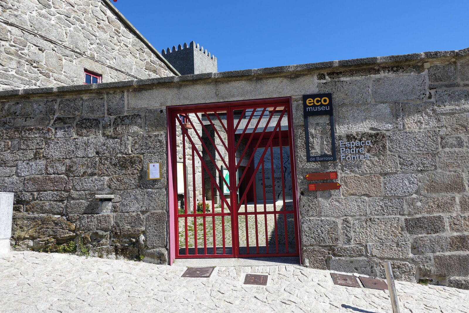Portão vermelho do Ecomuseu de Barroso, Espaço Padre Fontes, em muro de granito, com uma torre do castelo ao fundo